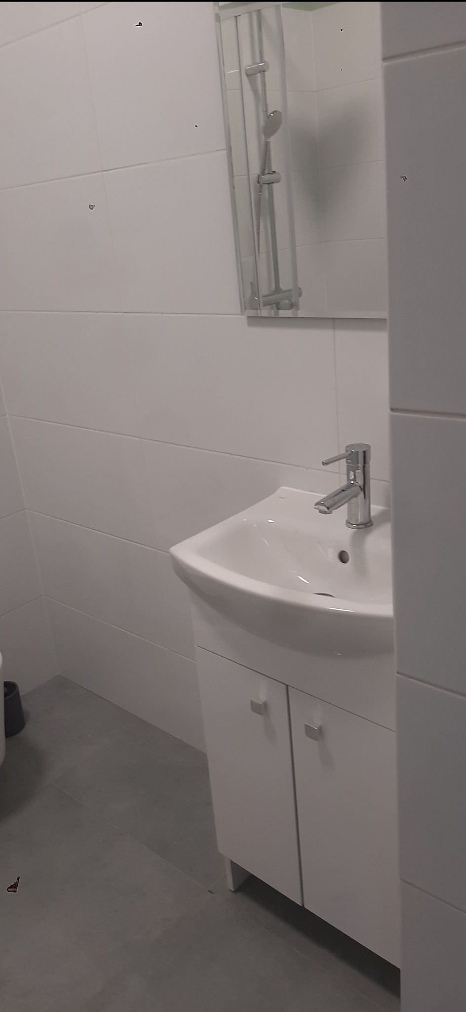 Biała umywalka z szafką w wyremontowanej łazience, widok na baterię i lustro z kabiną prysznicową. Szare płytki podłogowe, minimalistyczny styl.