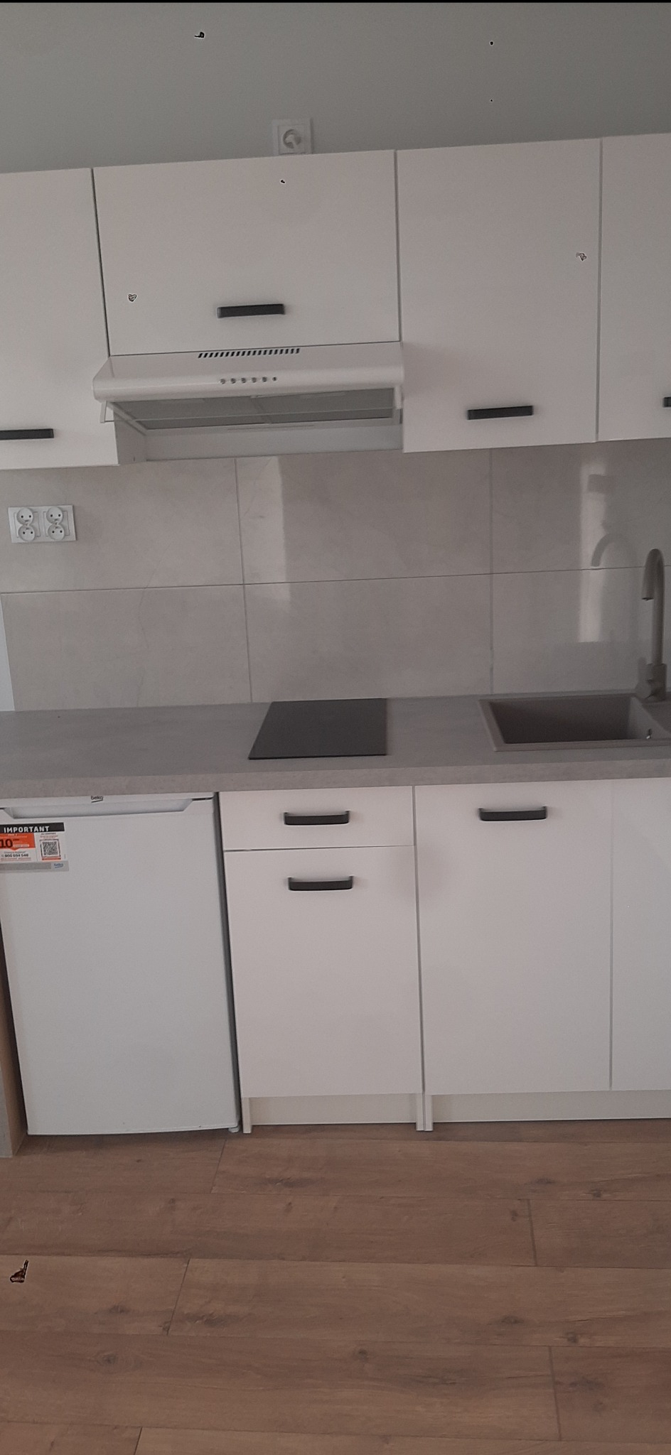 Biała zabudowa kuchenna z płytkami w odcieniach szarości, widoczny fragment podłogi z paneli. Minimalistyczny design, czarne uchwyty, płyta indukcyjna i mała lodówka.