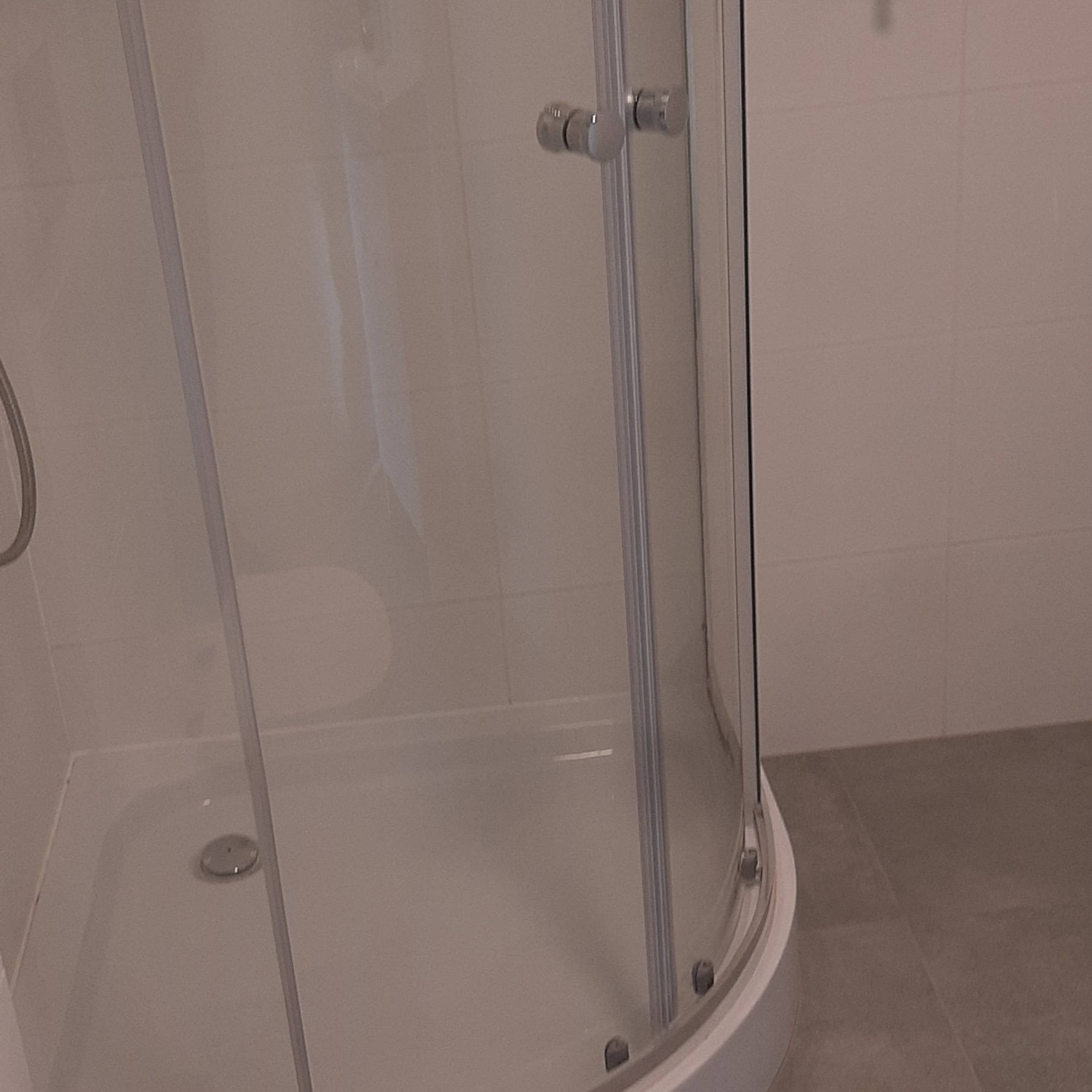 Szczegółowe ujęcie kabiny prysznicowej z przezroczystymi drzwiami i chromowanymi uchwytami, na tle jasnych płytek ściennych i szarej podłogi. Widoczny odpływ w brodziku.