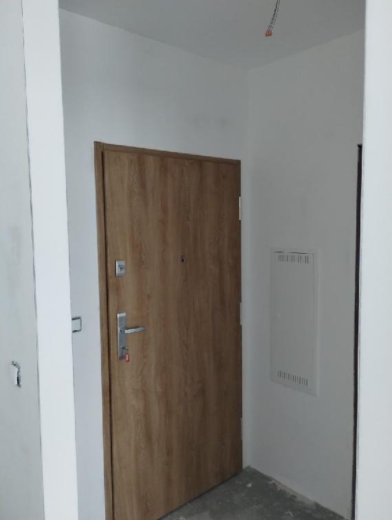 Nowe, drewnopodobne drzwi wejściowe w jasnym korytarzu, widoczne zamek i klamka. Na ścianie skrzynka elektryczna. Minimalistyczne wykończenie.