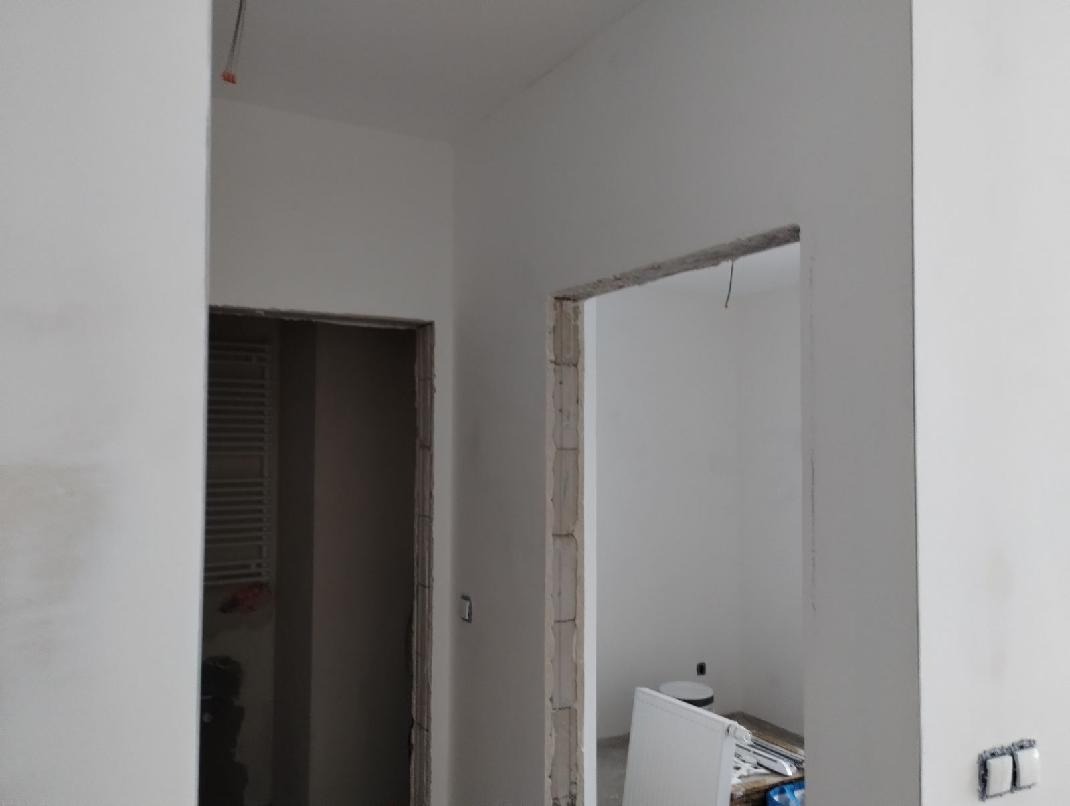 Wnętrze w trakcie prac wykończeniowych: widoczne surowe ściany, przygotowane otwory drzwiowe i fragment grzejnika. Minimalistyczny styl, jasne kolory.