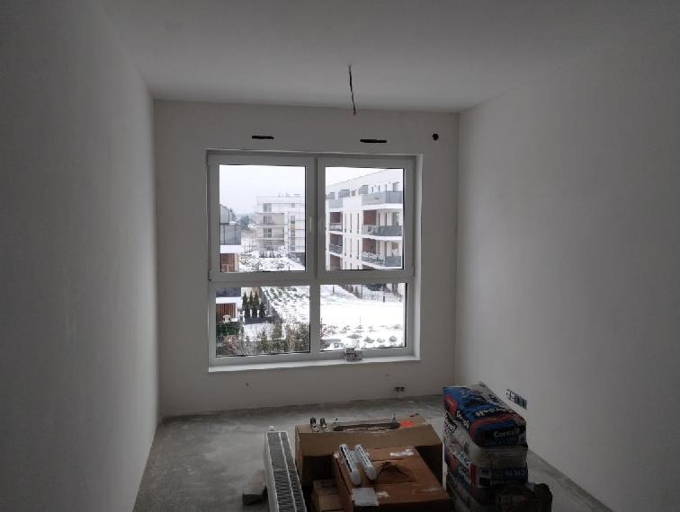 Wnętrze pokoju w trakcie remontu: gładkie, białe ściany, okno z widokiem na osiedle, worki z zaprawą i grzejnik. Przygotowanie do malowania lub wykończenia.