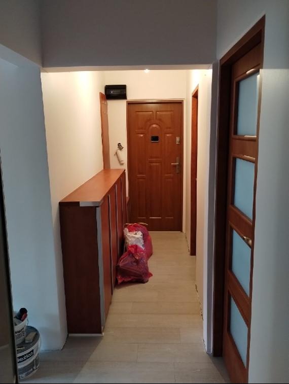 Wykończony korytarz z drewnianą szafką, drzwiami i panelami podłogowymi w odcieniach beżu. Widoczny worek z materiałami budowlanymi i wiadro z farbą.