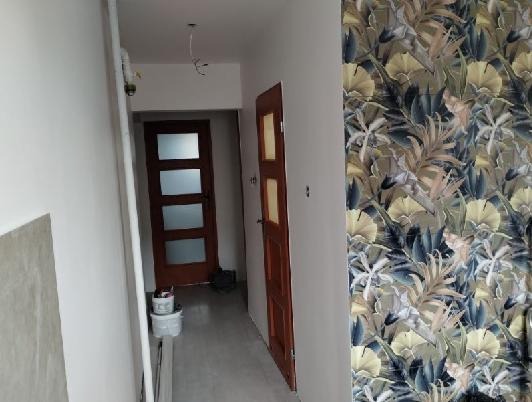 Wnętrze w trakcie prac: ściana z tapetą w roślinny wzór, widoczne drzwi wewnętrzne i przygotowane podłoże pod gładzie. Częściowo wykończone pomieszczenie.