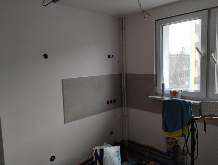 Fragment ściany w trakcie prac przygotowawczych do wykończenia, widoczne otwory pod instalacje i częściowo położona glazura w szarym odcieniu.
