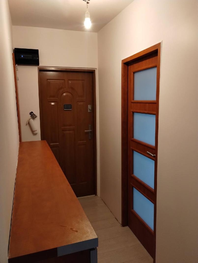 Jasne wnętrze przedpokoju z drewnianą komodą, drzwiami wejściowymi i wewnętrznymi z przeszkleniami. Świeże, białe ściany i subtelne oświetlenie.