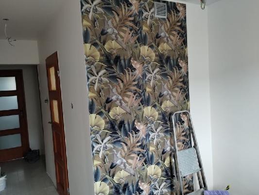 Ściana z tapetą w tropikalne wzory, drabina oparta o ścianę, widok na otwarte drzwi i fragment pomieszczenia. Wykończenie wnętrza z motywem roślinnym.