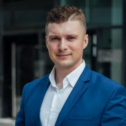 Portret mężczyzny w niebieskiej marynarce i białej koszuli, na tle nowoczesnego budynku w Piasecznie. Profesjonalny wygląd, biznesowy charakter.