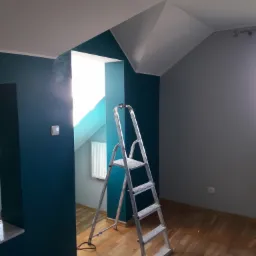 Wnętrze pokoju w trakcie odświeżania: ściany pomalowane na dwa kolory - głęboki turkus i szary, aluminiowa drabina oparta o ścianę, drewniana podłoga.