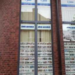 Wystawa agencji nieruchomości EuroDom z ogłoszeniami ofert sprzedaży mieszkań i domów, widoczna z zewnątrz przez duże okna witrynowe na tle ceglanej ściany budynku.