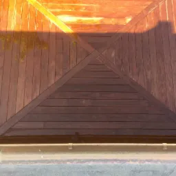 Drewniana konstrukcja z belek ułożonych w geometryczny wzór, oświetlona ciepłym światłem słonecznym, widok z góry na fragment tarasu z widocznym fragmentem rynny i betonowej powierzchni.