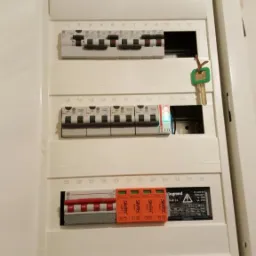 Otwarta skrzynka elektryczna z bezpiecznikami i wyłącznikami różnicowoprądowymi marki Legrand i Smitec, klucz do skrzynki w zamku.