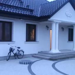 Biały dom z ciemną dachówką i kolumnami przy wejściu. Na pierwszym planie rower i ozdobny chodnik z kostki brukowej ułożonej w okrągły wzór.