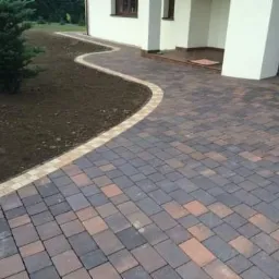 Ułożona z kostki brukowej ścieżka w ogrodzie, prowadząca do domu z brązowymi drzwiami garażowymi. Kostka w odcieniach brązu i szarości, obrzeże z jaśniejszych kostek. Po lewej stronie rabata...