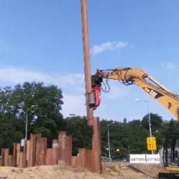 Żółta koparka CAT z czerwonym osprzętem wbija stalowy profil w ziemię, tworząc ścianę szczelinową; widoczne inne wbite profile i fragment drogi w tle.