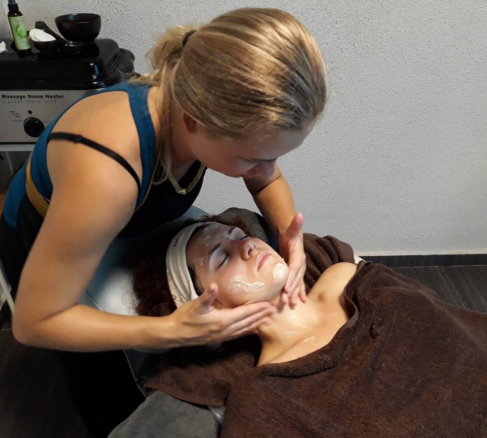 Kobieta wykonuje manualny zabieg na twarzy klientki leżącej na leżance, widoczny podgrzewacz do kamieni, krem na twarzy klientki.