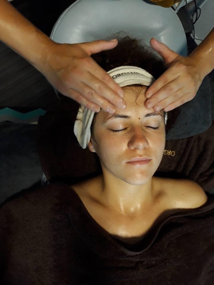 Kobieta z zamkniętymi oczami otrzymuje masaż twarzy; widoczne dłonie masażysty, opaska na włosach z napisem 'ORGANIC', skóra pokryta olejkiem.