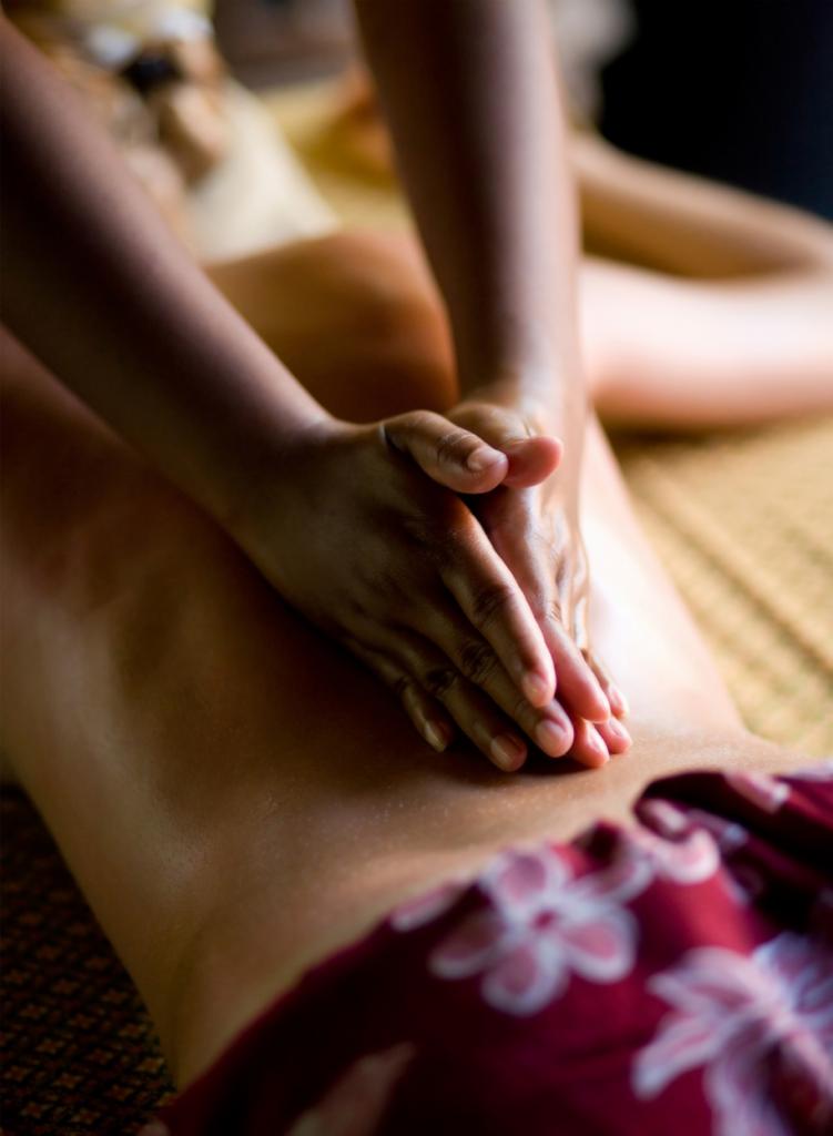 Dłonie masażysty masują plecy klienta leżącego na macie, widoczny fragment kwiecistego pareo.