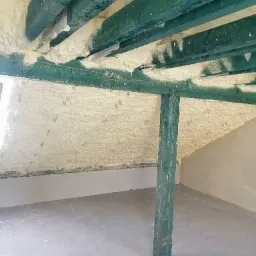 Poddasze z drewnianą konstrukcją pokryte jasną pianą izolacyjną, widoczny fragment ściany i podłogi, pionowy słup konstrukcyjny pomalowany na zielono.