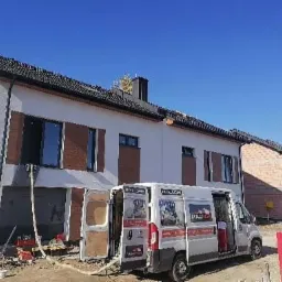 Dwukondygnacyjny dom w trakcie prac izolacyjnych, z widocznym wężem doprowadzającym materiał i samochodem dostawczym z otwartymi drzwiami, na tle błękitnego nieba.