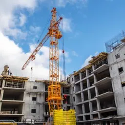 Wysoki żuraw budowlany w kolorze pomarańczowym na tle częściowo wzniesionego, wielopiętrowego budynku z betonu, widoczne szalunki i zbrojenia.