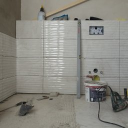 Wett - Proces układania płytek o falistej strukturze w łazience, widoczny poziomica, puszka instalacyjna i narzędzia.