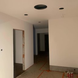 Wett - Wnętrze w trakcie prac wykończeniowych: świeżo pomalowane na biało ściany, widoczne otwory w suficie pod oświetlenie, zabezpieczona podłoga, częściowo obrobione ościeżnice drzwiowe, puszki...