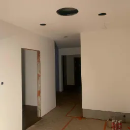 Wnętrze w trakcie prac wykończeniowych: świeżo pomalowane na biało ściany, widoczne otwory w suficie pod oświetlenie, zabezpieczona podłoga, częściowo obrobione ościeżnice drzwiowe, puszki...