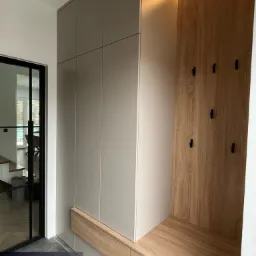 Wnęka z szafami w odcieniach szarości i siedziskiem z drewnopodobną okładziną, oświetlona ledowym paskiem, z czarnymi haczykami na ścianie, widoczna z perspektywy wejścia do pomieszczenia.