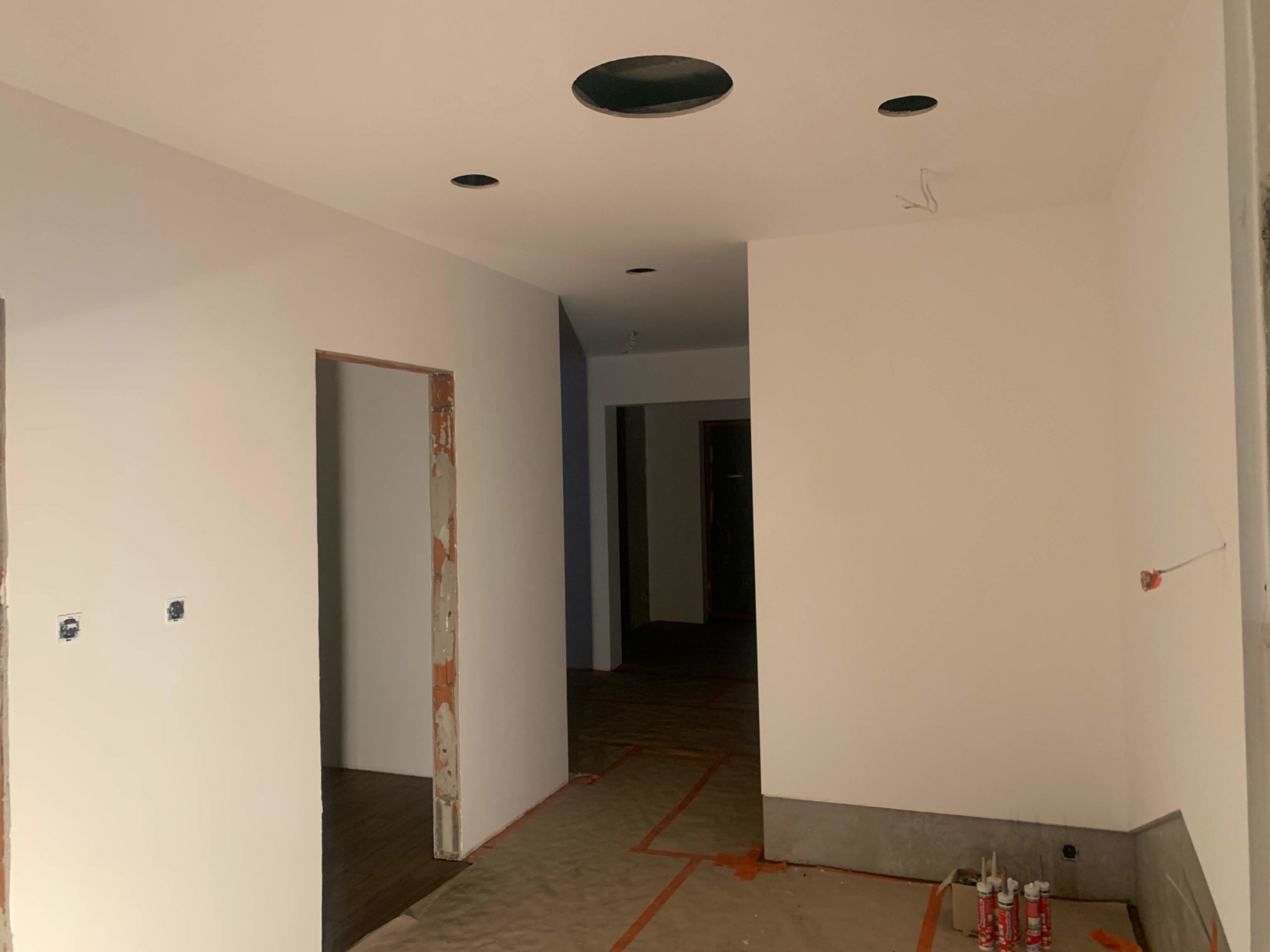 Wnętrze w trakcie prac wykończeniowych: świeżo pomalowane na biało ściany, widoczne otwory w suficie pod oświetlenie, zabezpieczona podłoga, częściowo obrobione ościeżnice drzwiowe, puszki...