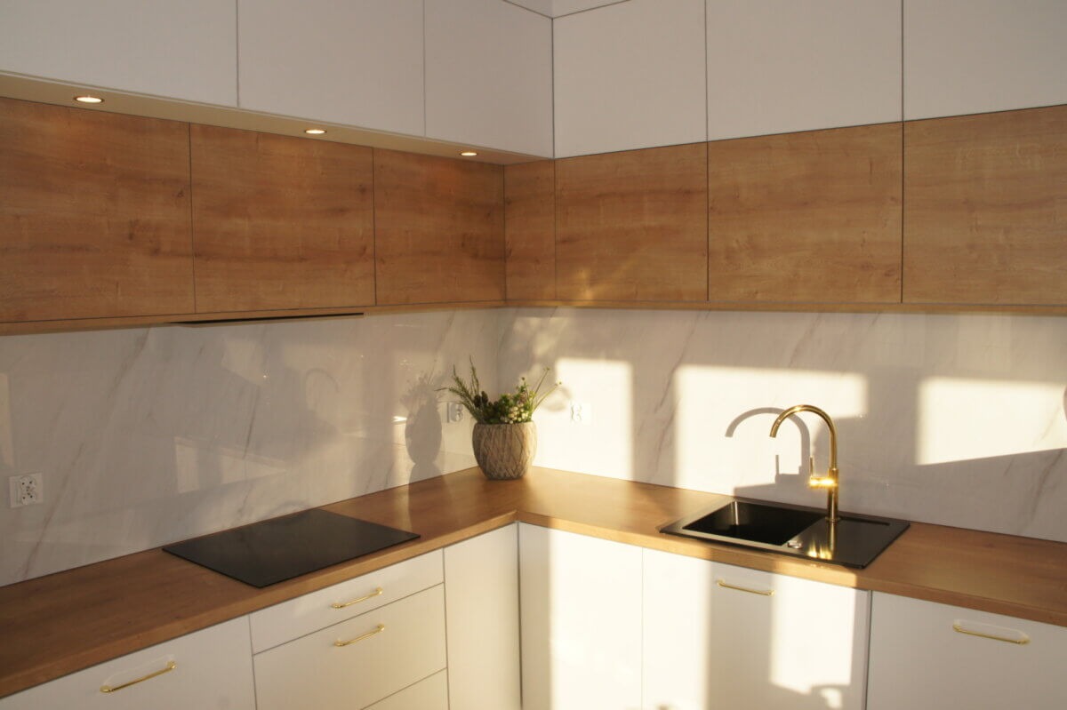 Nowoczesna kuchnia z białymi szafkami dolnymi i drewnopodobnymi górnymi, marmurowy backsplash, złota bateria i czarny zlew, oświetlenie punktowe pod szafkami górnymi, na blacie płyta indukcyjna...