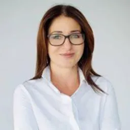 Portret kobiety w okularach i białej koszuli na jasnoszarym tle. Uśmiech, profesjonalny wygląd.