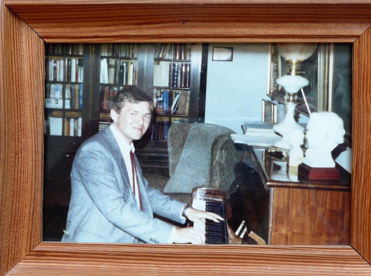 Oprawione zdjęcie mężczyzny w garniturze grającego na pianinie w pokoju z biblioteczką i biurkiem z lampą i popiersiami.