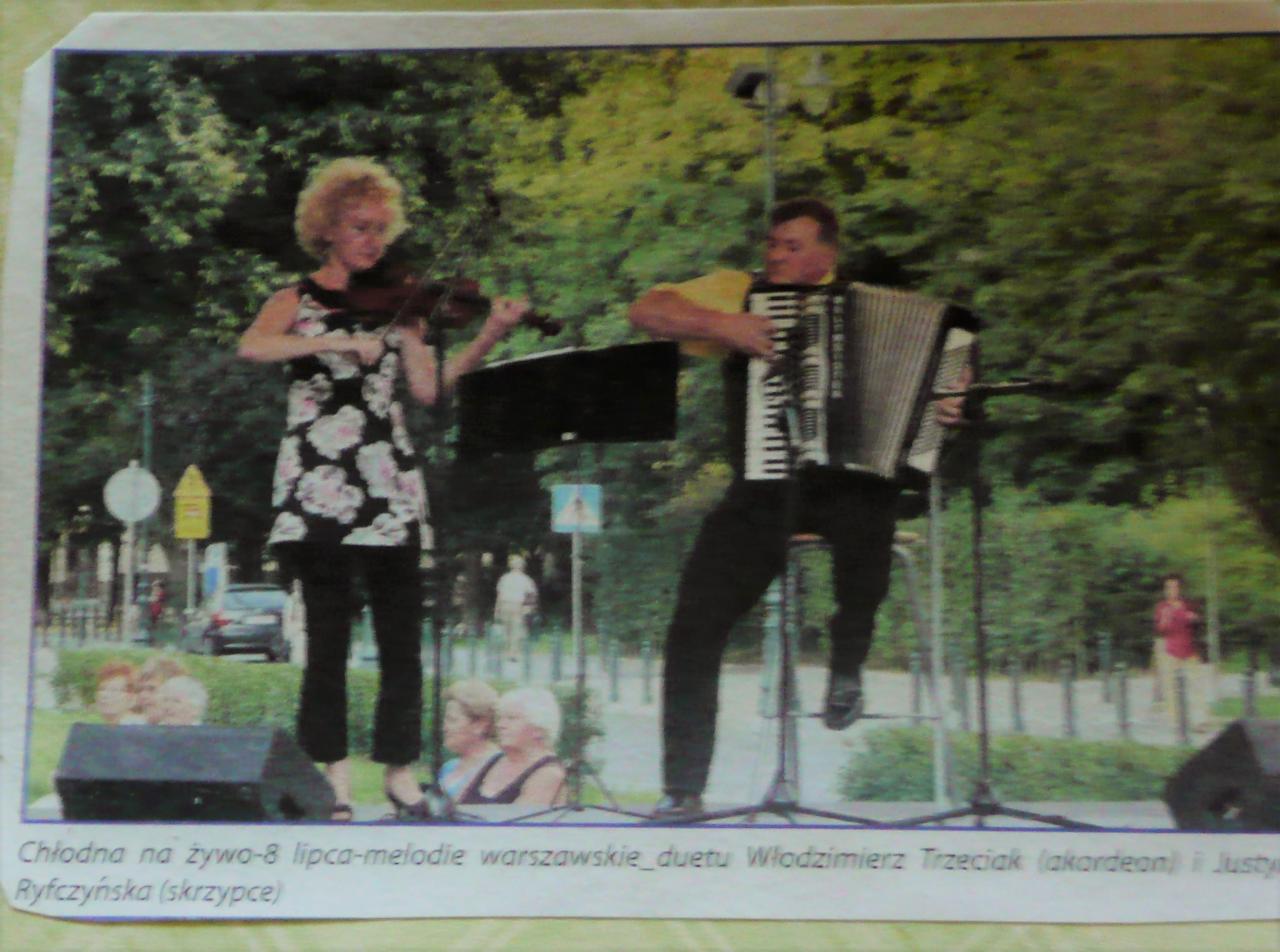 Duet muzyczny na scenie plenerowej: kobieta gra na skrzypcach w kwiecistej tunice, mężczyzna na akordeonie, z widoczną publicznością i zielenią w tle.