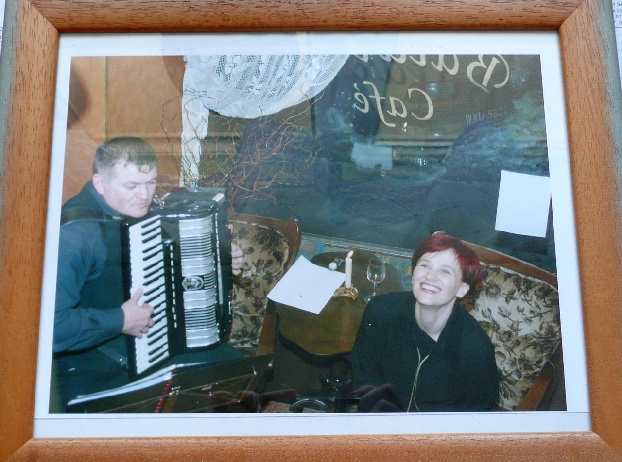 Zdjęcie w ramce przedstawia mężczyznę grającego na akordeonie oraz uśmiechniętą kobietę siedzącą obok stolika z zapaloną świecą w tle szyby z napisem 'Cafe'.