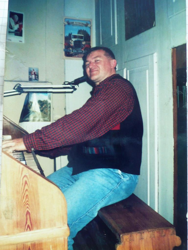 Mężczyzna w koszuli w kratę gra na drewnianych organach w pomieszczeniu z plakatami na ścianie.