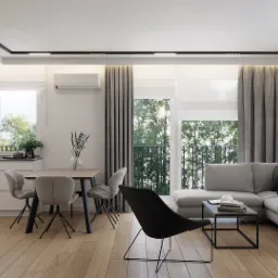 Jasne, przestronne wnętrze mieszkania z drewnianą podłogą, szarą sofą narożną, stołem z krzesłami i czarnym fotelem na tle okien z widokiem na zieleń.