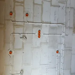 Świeżo położona instalacja elektryczna w ścianie z betonu komórkowego, widoczne białe kable w peszlach i pomarańczowe puszki instalacyjne, ściana z widocznymi oznaczeniami i notatkami wykonawcy.
