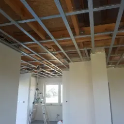 Montaż metalowego stelażu sufitowego w pomieszczeniu z surowymi ścianami, widoczna drabina i laserowy niwelator.