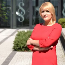 Portret kobiety w czerwonej sukience, z założonymi rękami, na tle budynku z symbolem paragrafu na szklanych drzwiach.