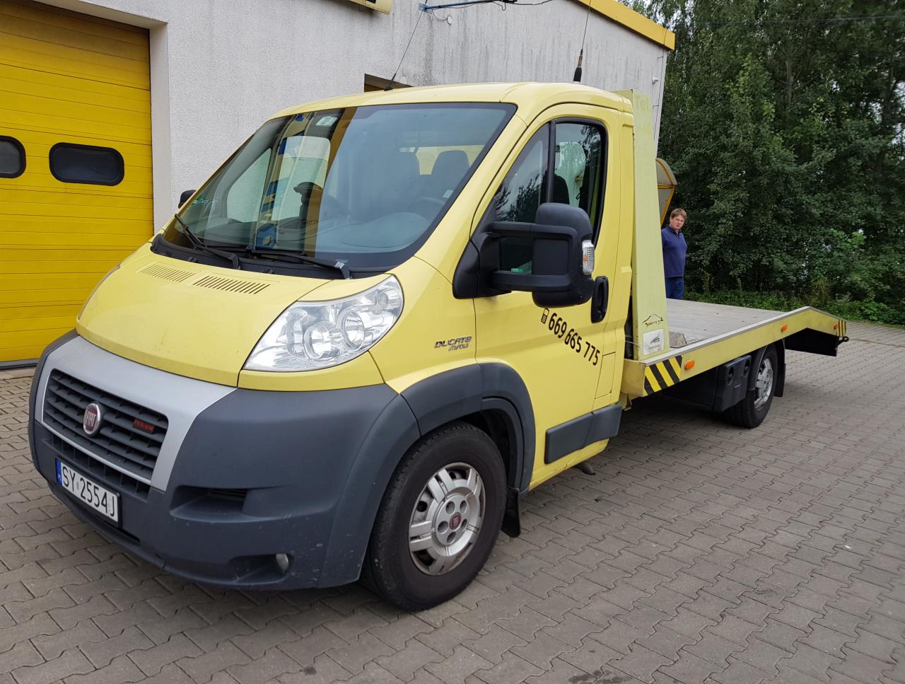 Żółta laweta Fiat Ducato z numerem telefonu na drzwiach, zaparkowana na kostce brukowej przed budynkiem z żółtą bramą garażową.