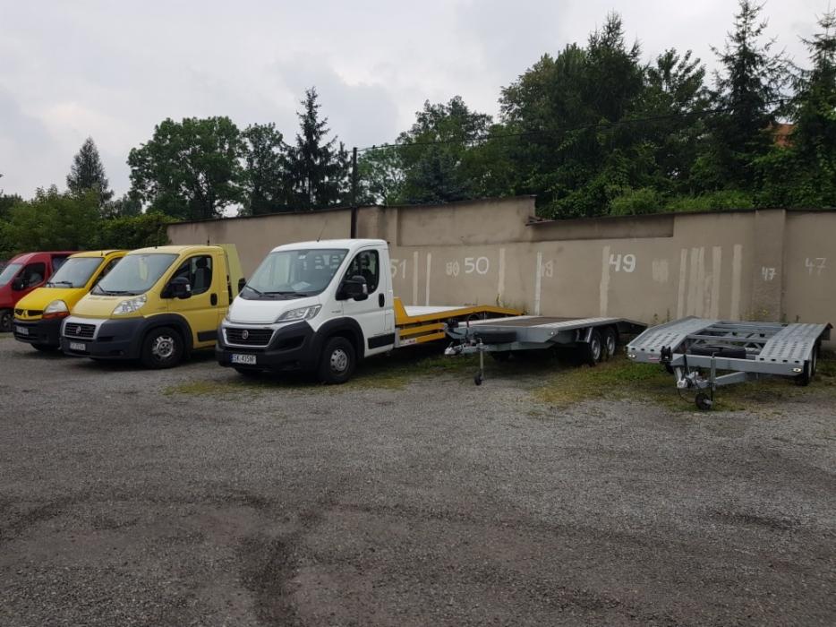 Flota lawet i samochodów transportowych na żwirowym parkingu, w tle betonowy mur z numeracją.