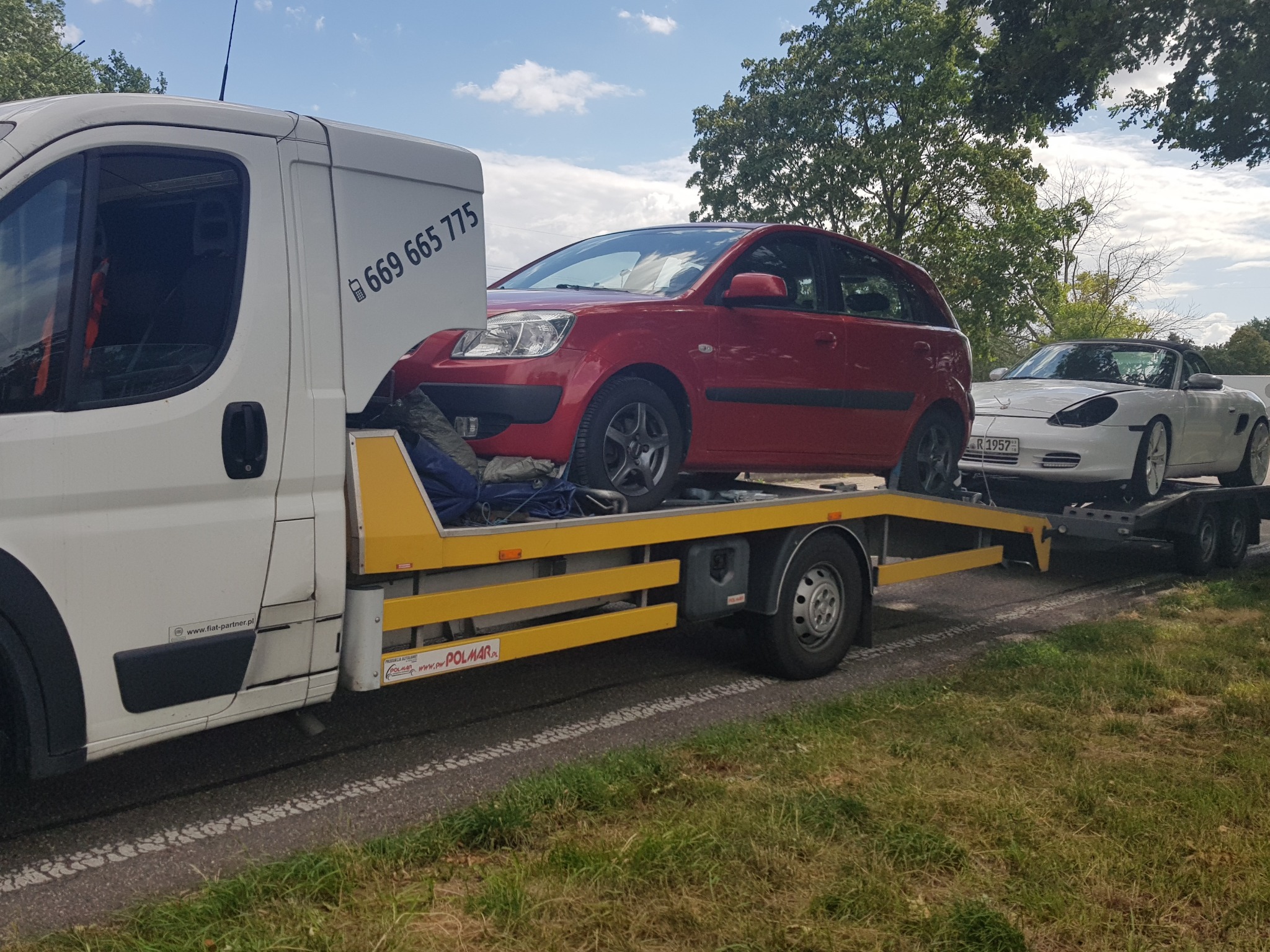 Laweta z czerwonym hatchbackiem i białym kabrioletem na platformie, widoczny numer telefonu na kabinie lawety.