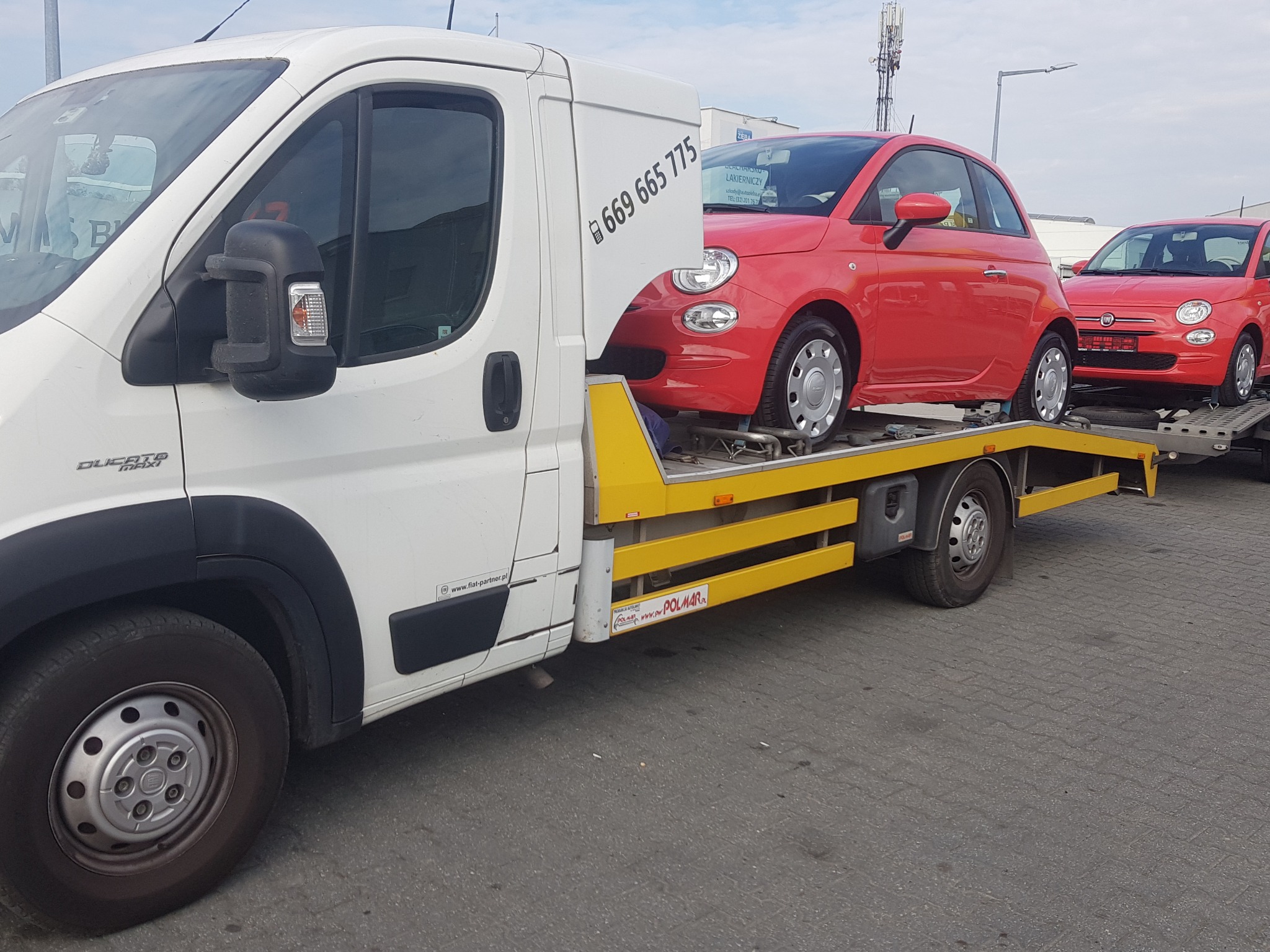 Biała laweta z żółtymi elementami transportuje dwa czerwone samochody Fiat 500 na parkingu w słoneczny dzień. Na zabudowie lawety widoczny numer telefonu.