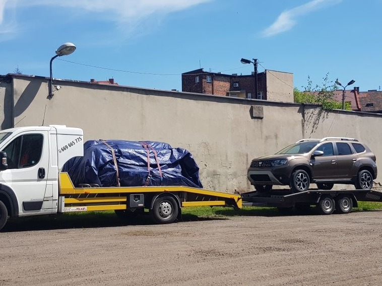 Laweta z brązowym SUVem Dacia Duster oraz ładunkiem zabezpieczonym niebieską plandeką na parkingu w słoneczny dzień.