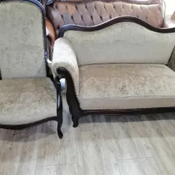 Odnowiony komplet mebli w stylu retro: fotel z wysokim oparciem i dwuosobowa sofa, obite jasną tkaniną w delikatny, roślinny wzór, z ciemnobrązowymi, giętymi nogami i ozdobnymi pinezkami.