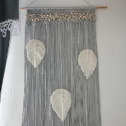 Szara makrama ścienna z trzema białymi piórami, zawieszona na drewnianym drążku z ozdobnymi drewnianymi kulkami.