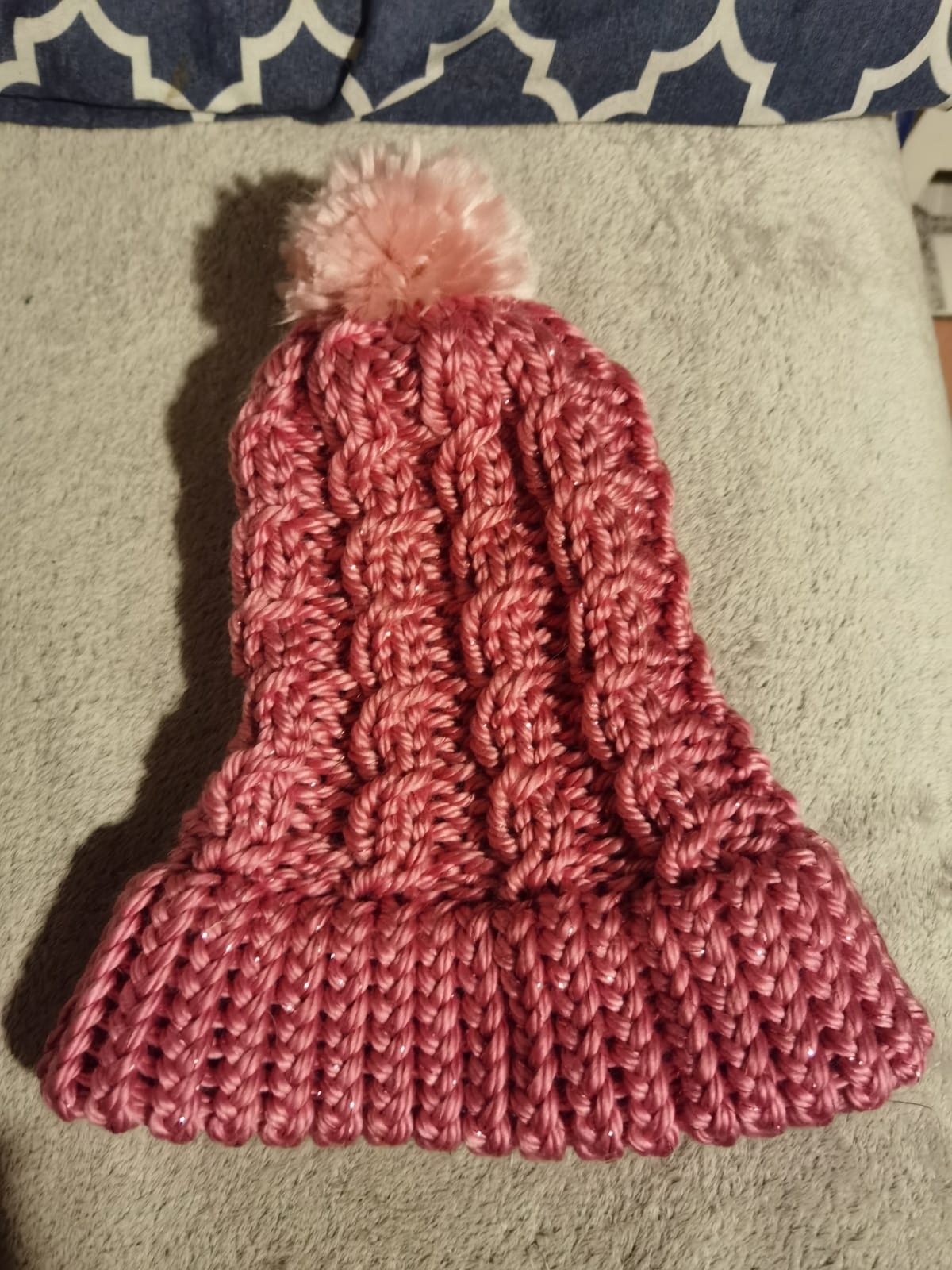 Ręcznie robiona, różowa czapka z pomponem, wykonana na drutach z błyszczącej włóczki, leżąca na szarym, pluszowym tle.
