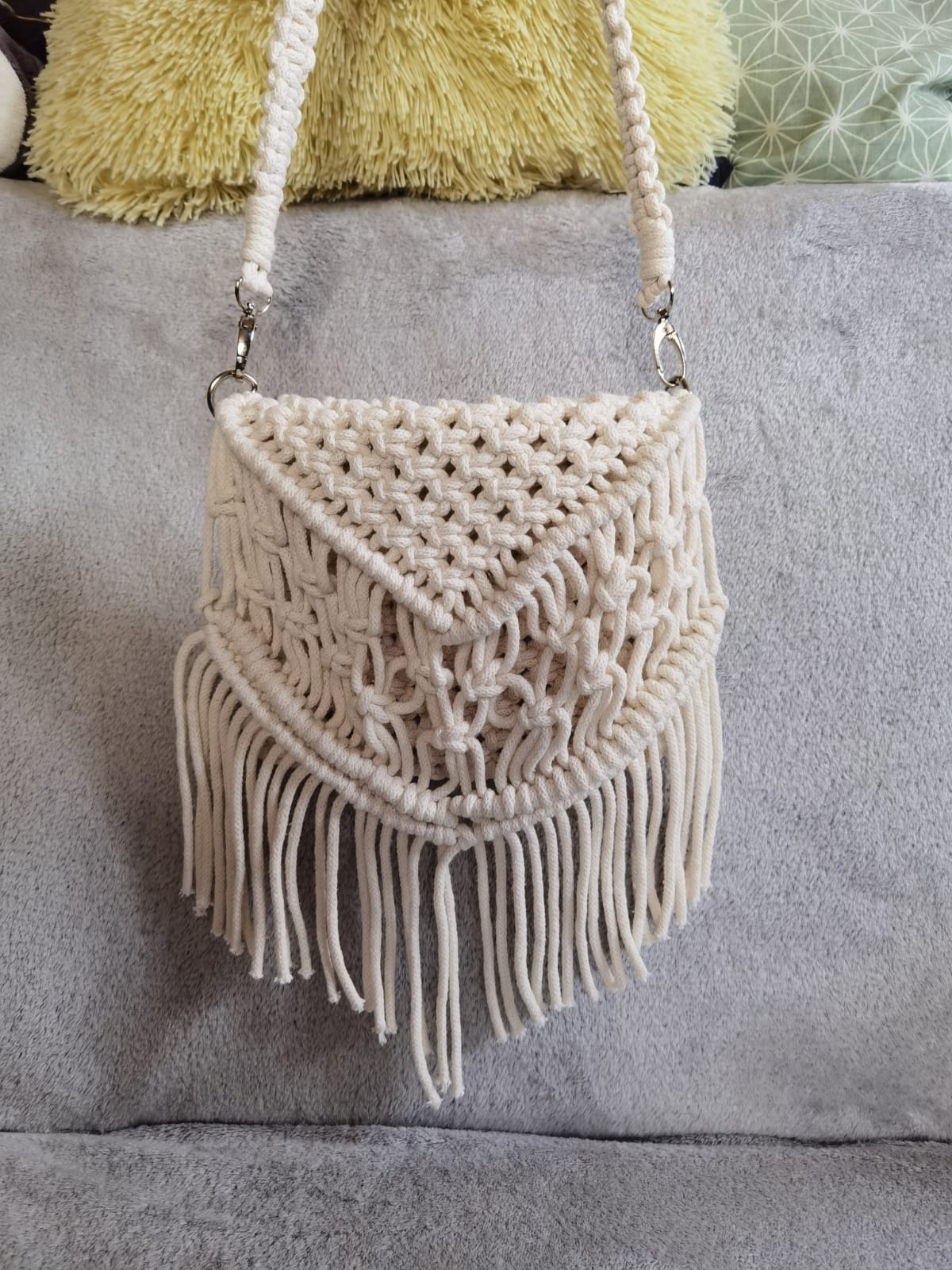 Ręcznie robiona torebka z makramy w odcieniach bieli, wisząca na szarym tle, z widocznymi detalami splotu i frędzli.