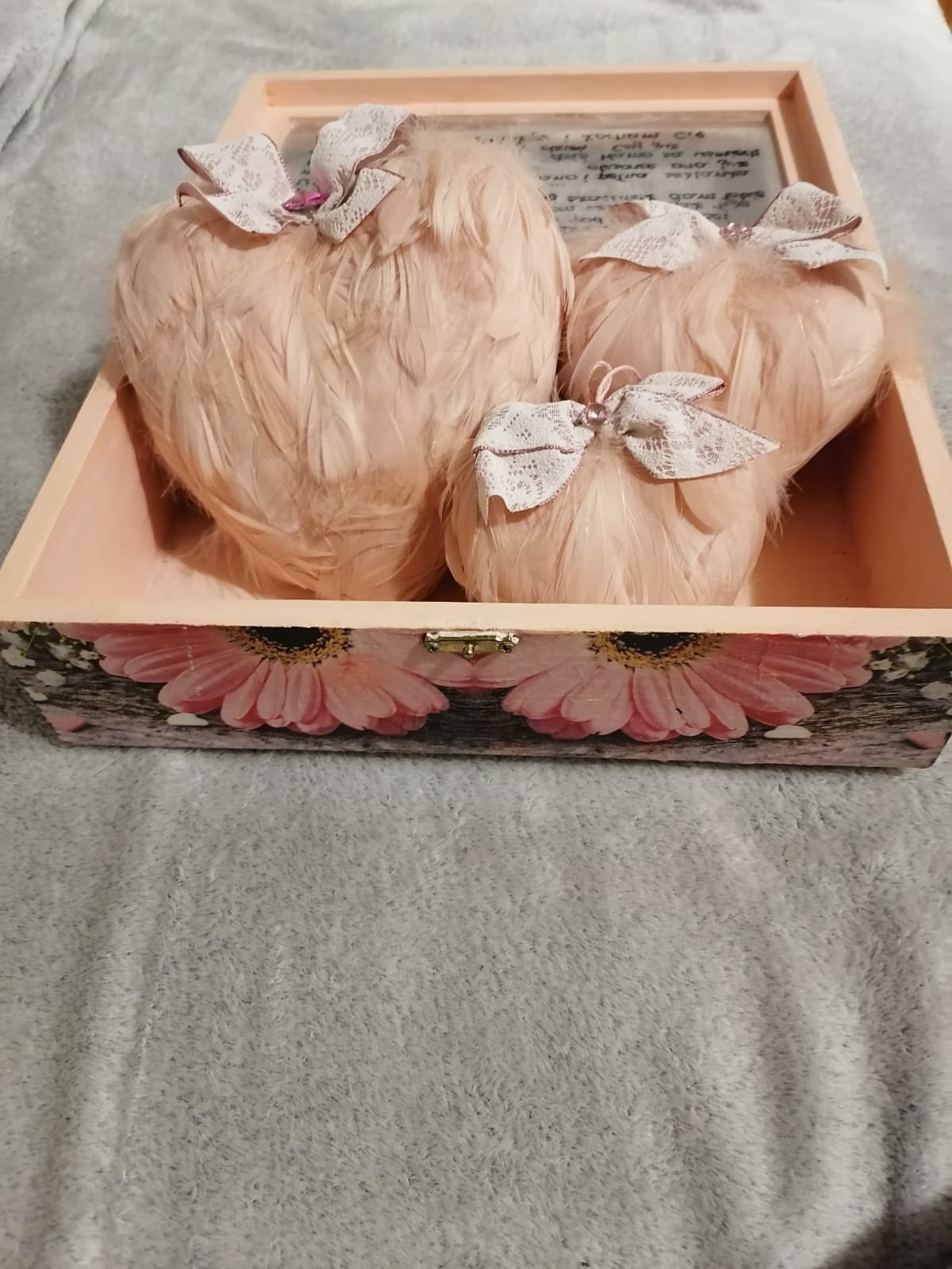 Ręcznie robione, różowe serca z piórkami i koronkowymi kokardkami w drewnianej szkatułce z motywem kwiatowym, leżącej na szarym kocu.